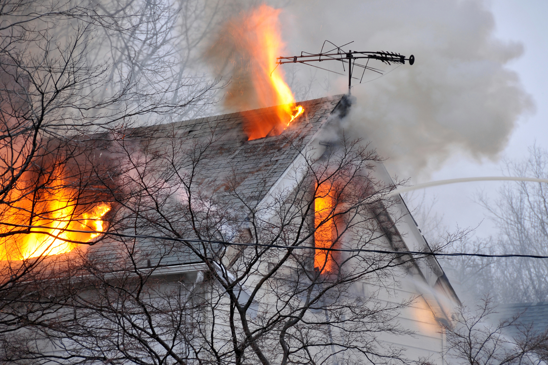 House on fire, flames and smoke billowing from the roof and windows. Bare winter trees in the foreground.