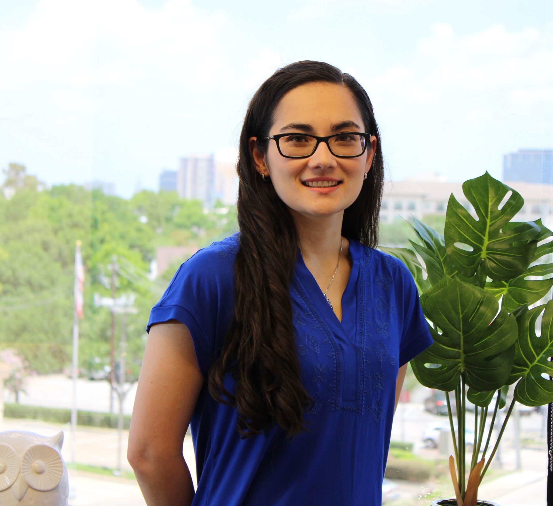 A woman in a blue shirt stands in front of a window with flowers in a vase