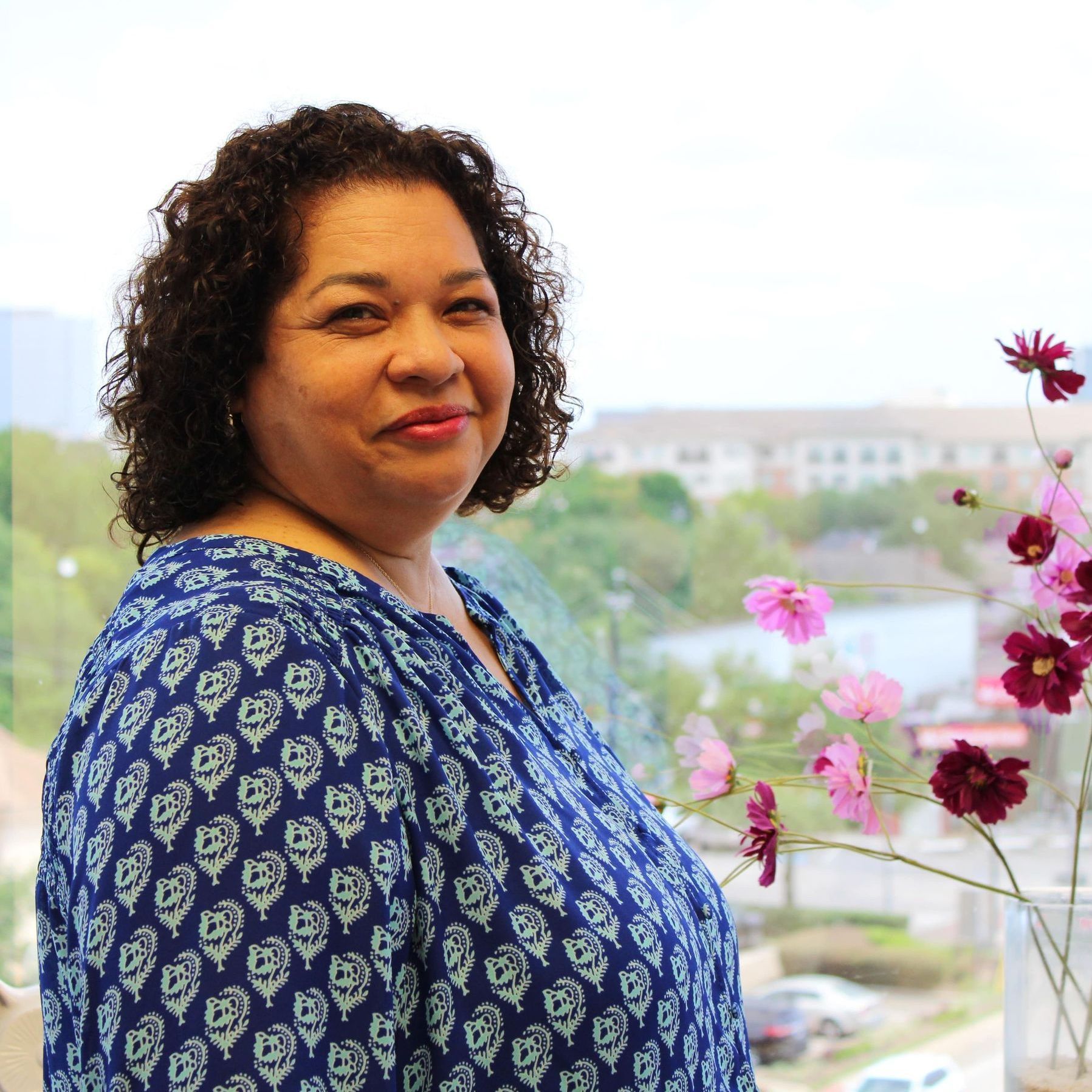 A woman in a blue shirt stands in front of a window with flowers in a vase