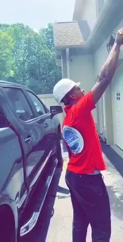 A man in a red shirt is standing next to a black truck in front of a house.