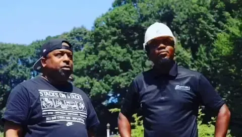 Two men are standing next to each other in a park wearing hard hats.