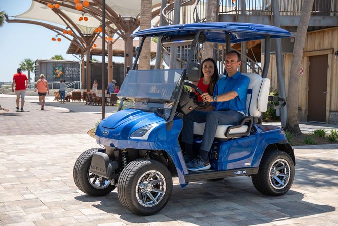 Couple in blue golf cart smiles as they drive on a paved area with people and buildings visible in the background.