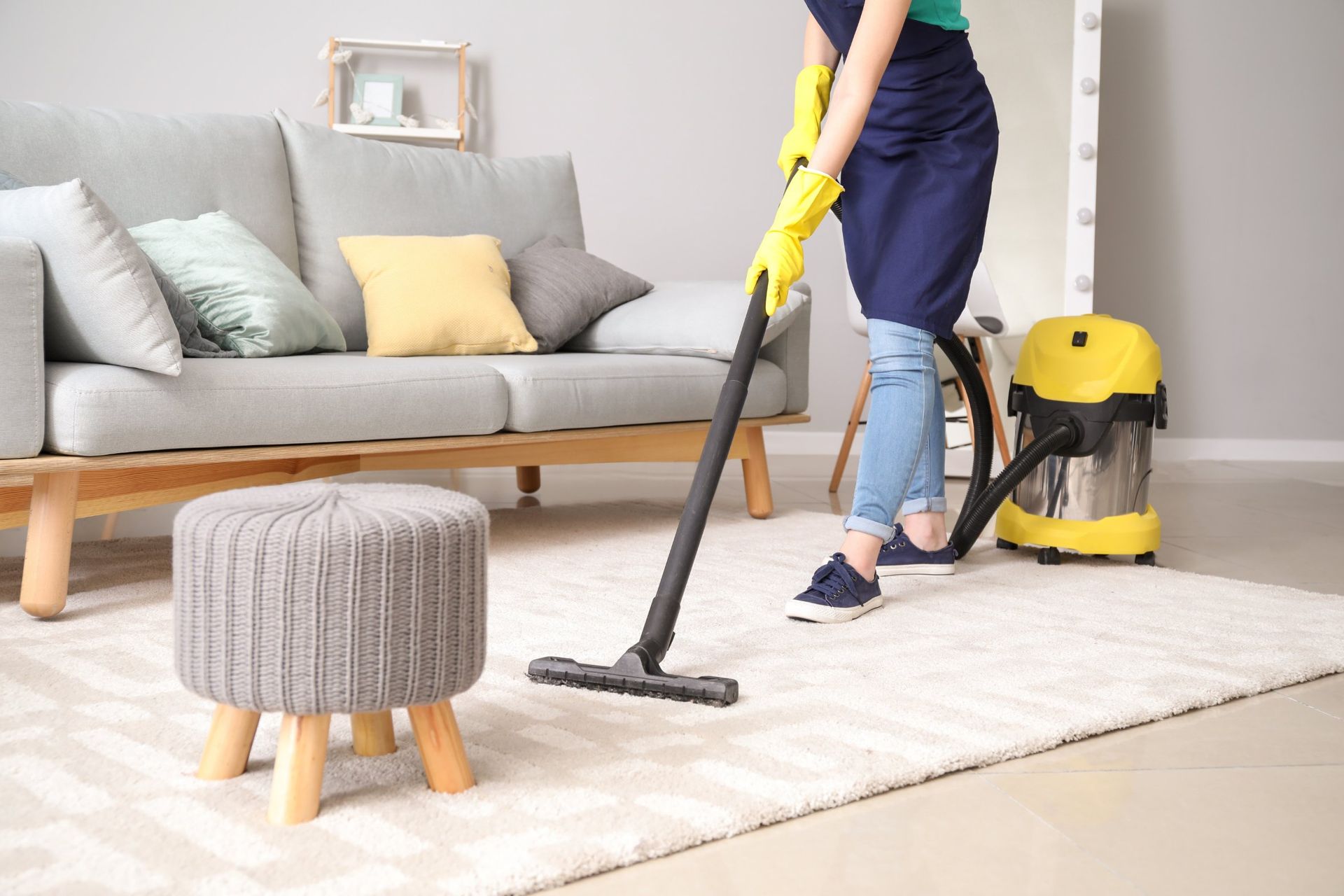 Person vacuums a white rug in a living room, wearing gloves and an apron, next to a sofa and a stool.