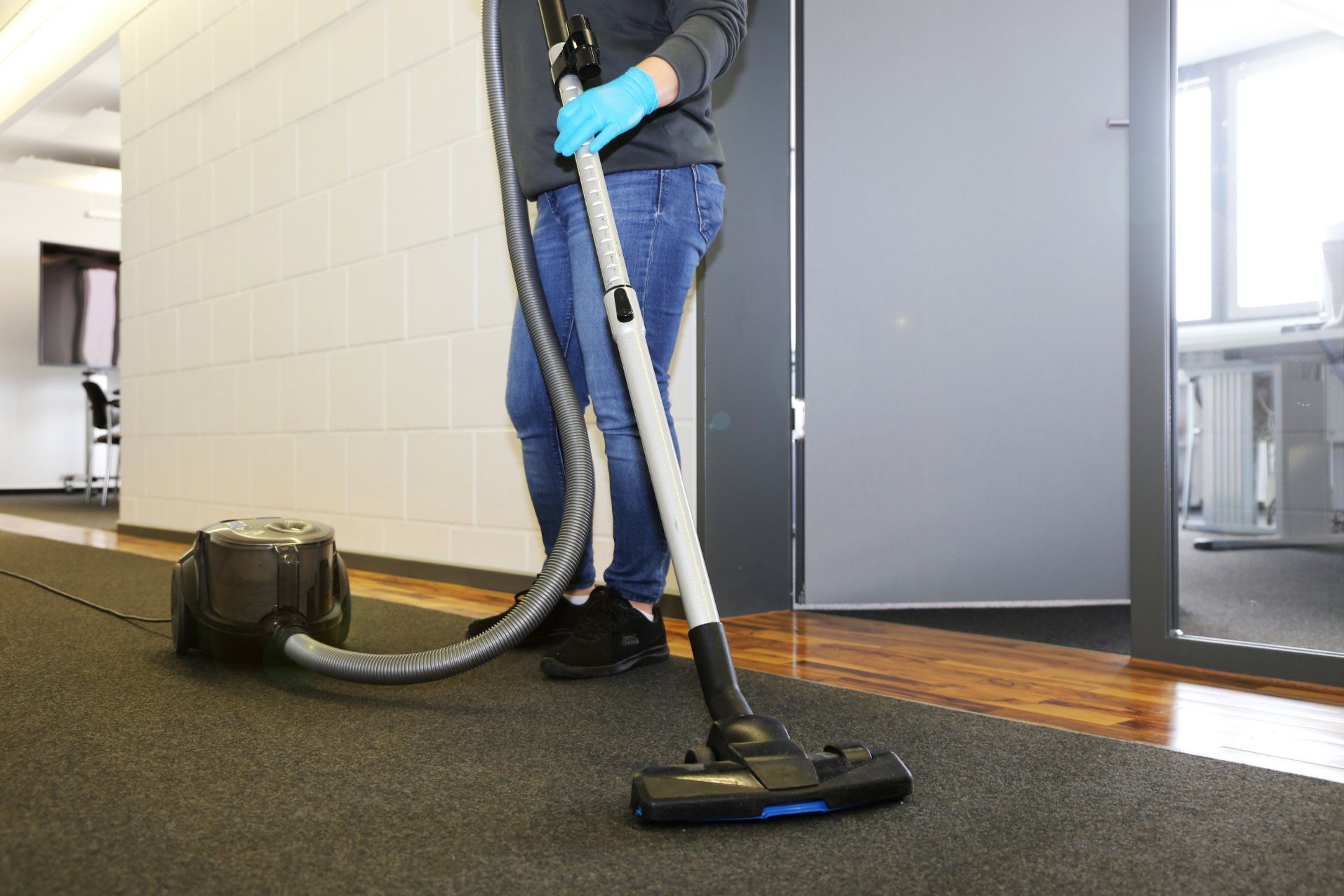 Person vacuuming a dark carpeted floor in an office. They are wearing blue gloves and jeans.