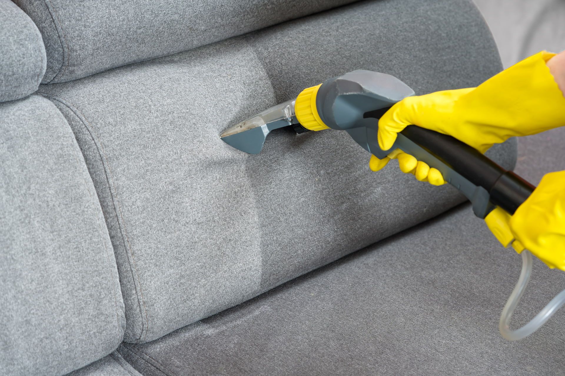 Person in yellow gloves cleaning a gray sofa with a steam cleaner.