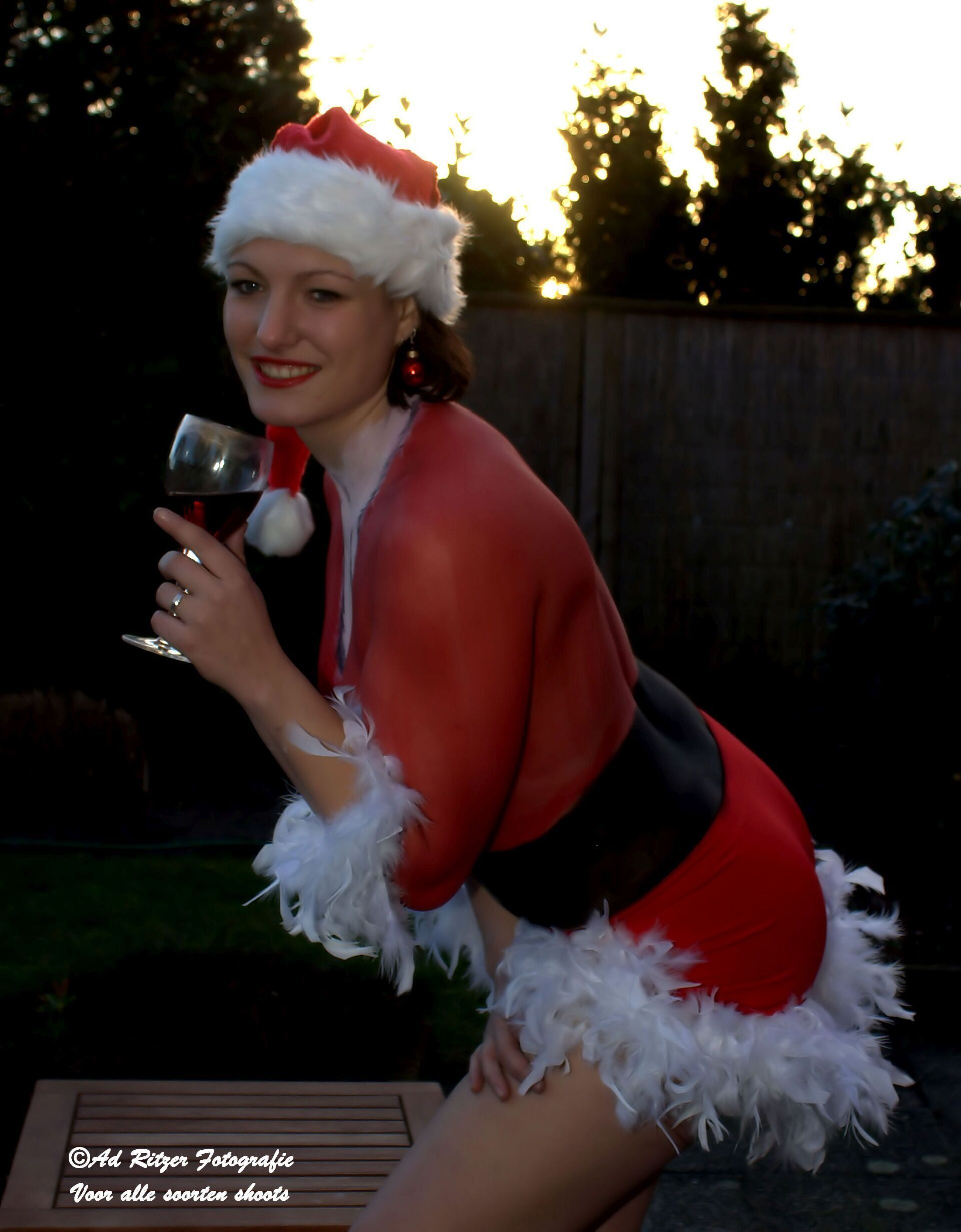 Vrouw in een kerstmanpak poseert buiten met een wijnglas bij zonsondergang.