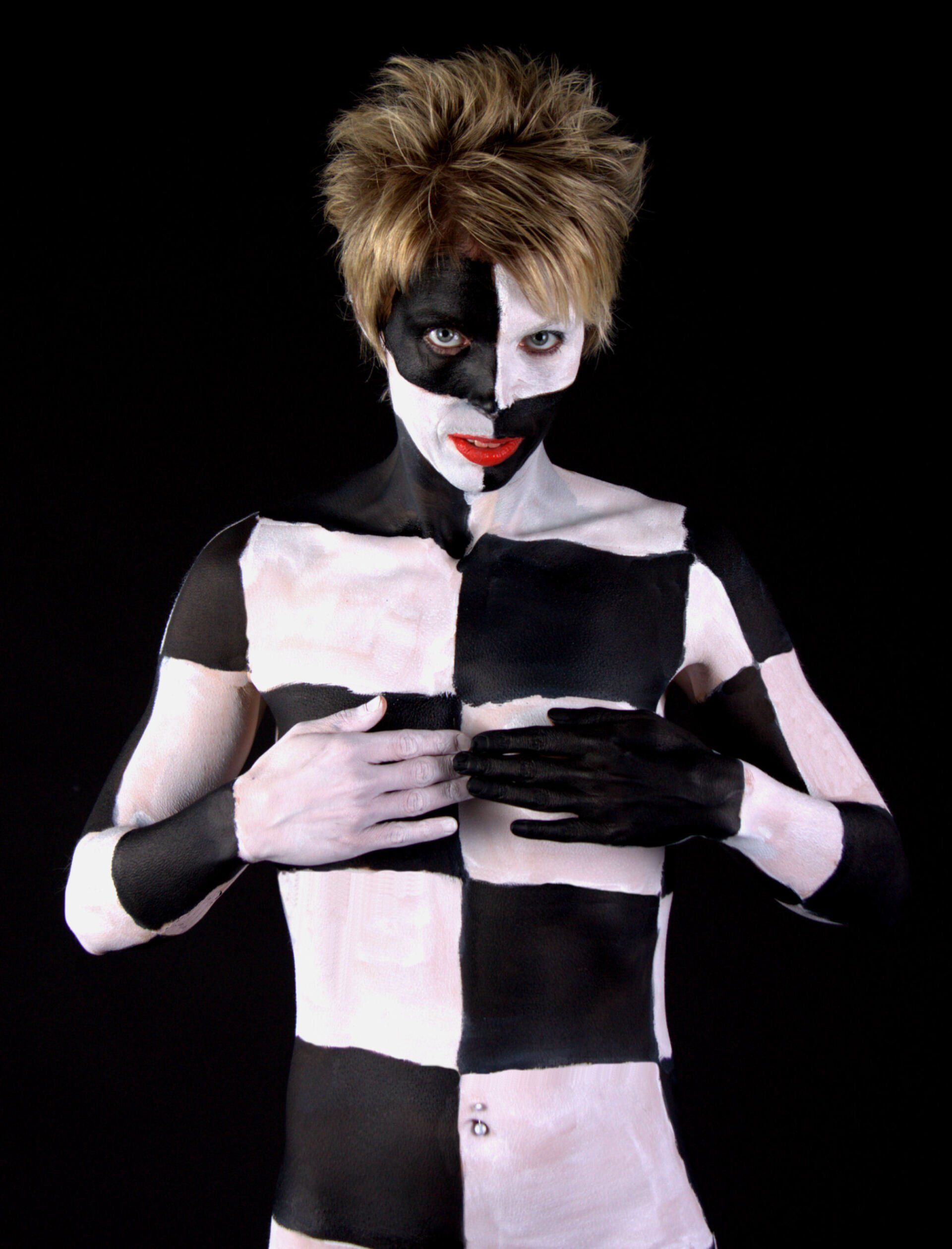 Persoon met zwart-witte bodypaint en stekelig haar, kijkt naar voren tegen een donkere achtergrond.