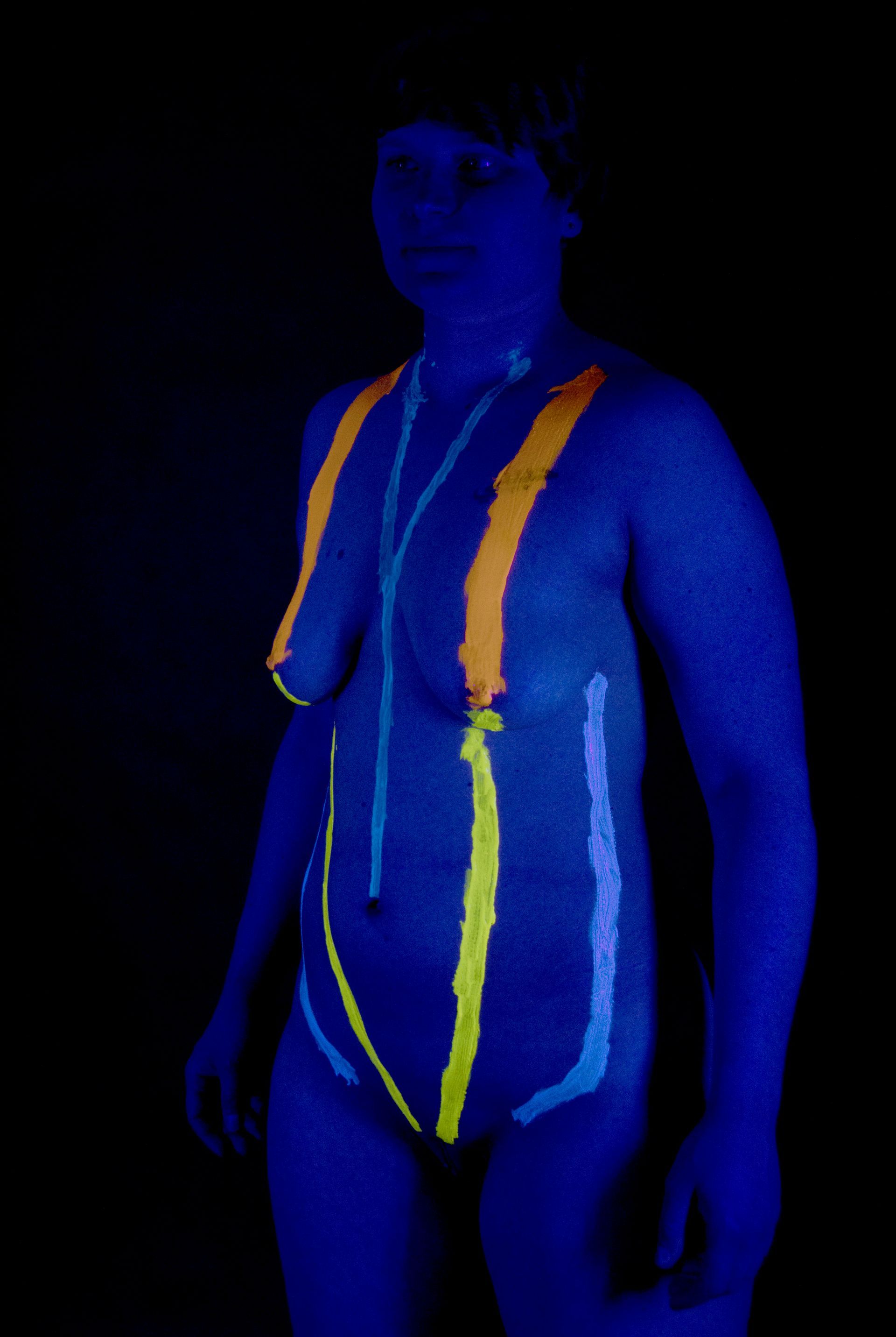 Persoon met blauwe lichaamsverf en felgele/oranje markeringen, staand tegen een zwarte achtergrond.