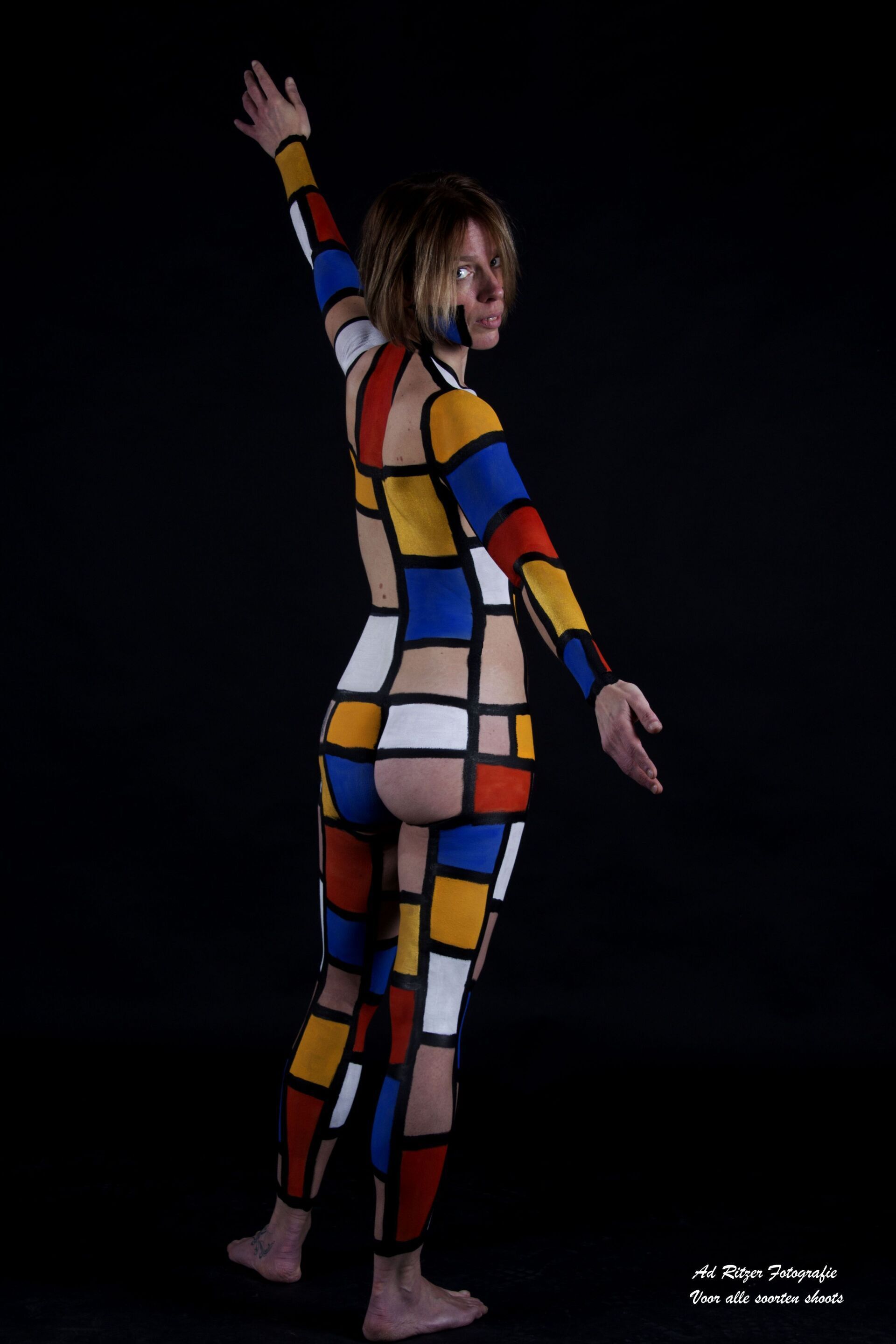 Persoon in een kleurrijk geometrisch bodysuit poseert tegen een zwarte achtergrond, met één arm omhoog.