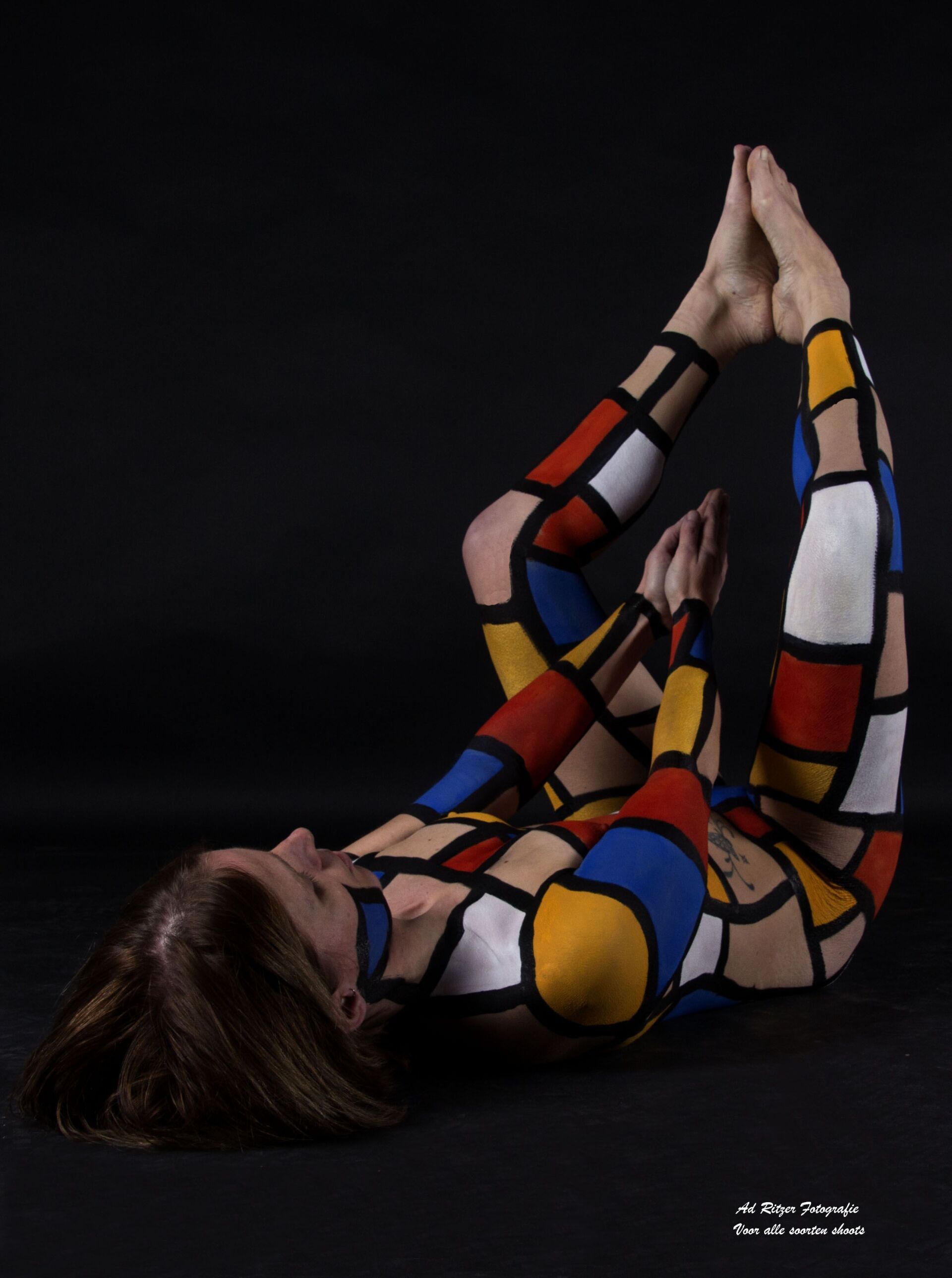 Persoon ligt op een zwarte achtergrond, benen omhoog, lichaam beschilderd met opvallende rode, blauwe, gele en zwarte geometrische blokken.