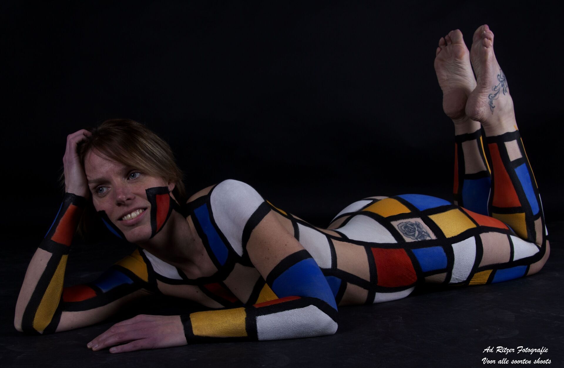 Vrouw in een kleurrijk geometrisch bodysuit poseert op een donkere studiovloer, glimlachend met haar voeten omhoog achter zich.