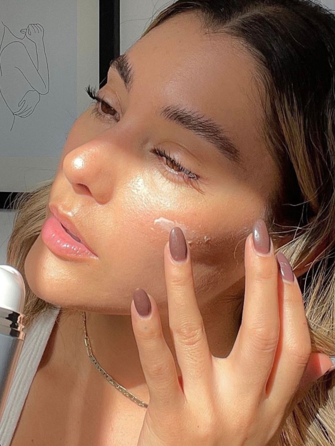 Close-up of a woman applying cream to her cheek in warm sunlight, with manicured nails and soft makeup