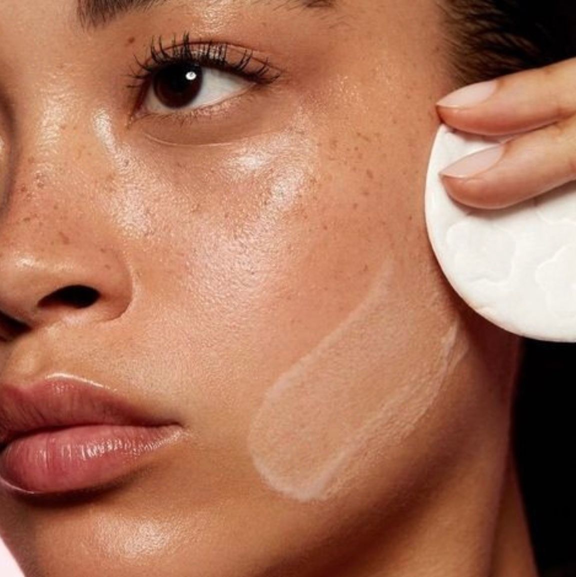 Close-up of a woman applying white skincare cream to her cheek with a cotton pad