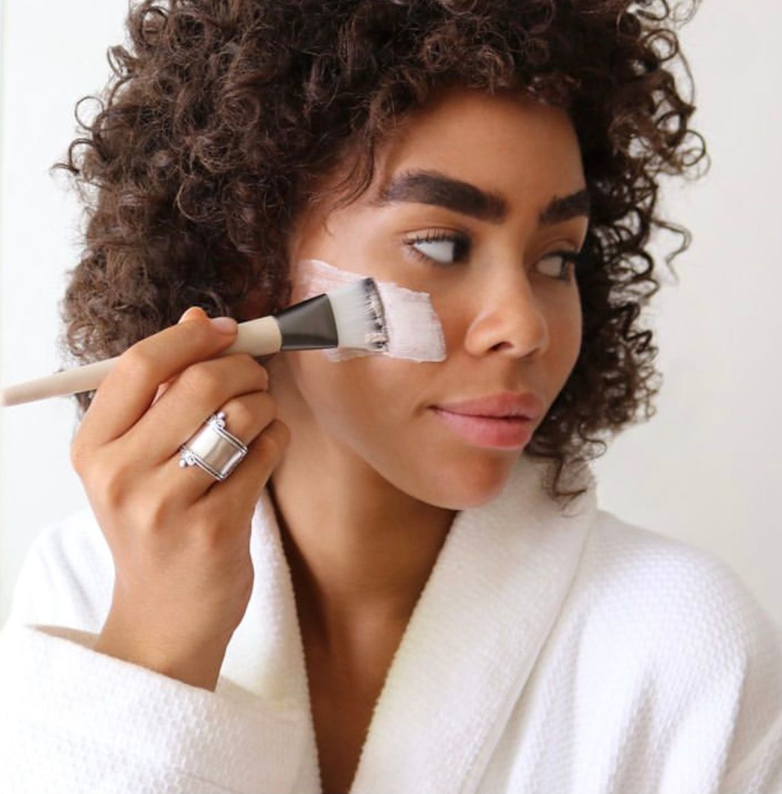 Woman applying a facial mask with a brush while wearing a white robe