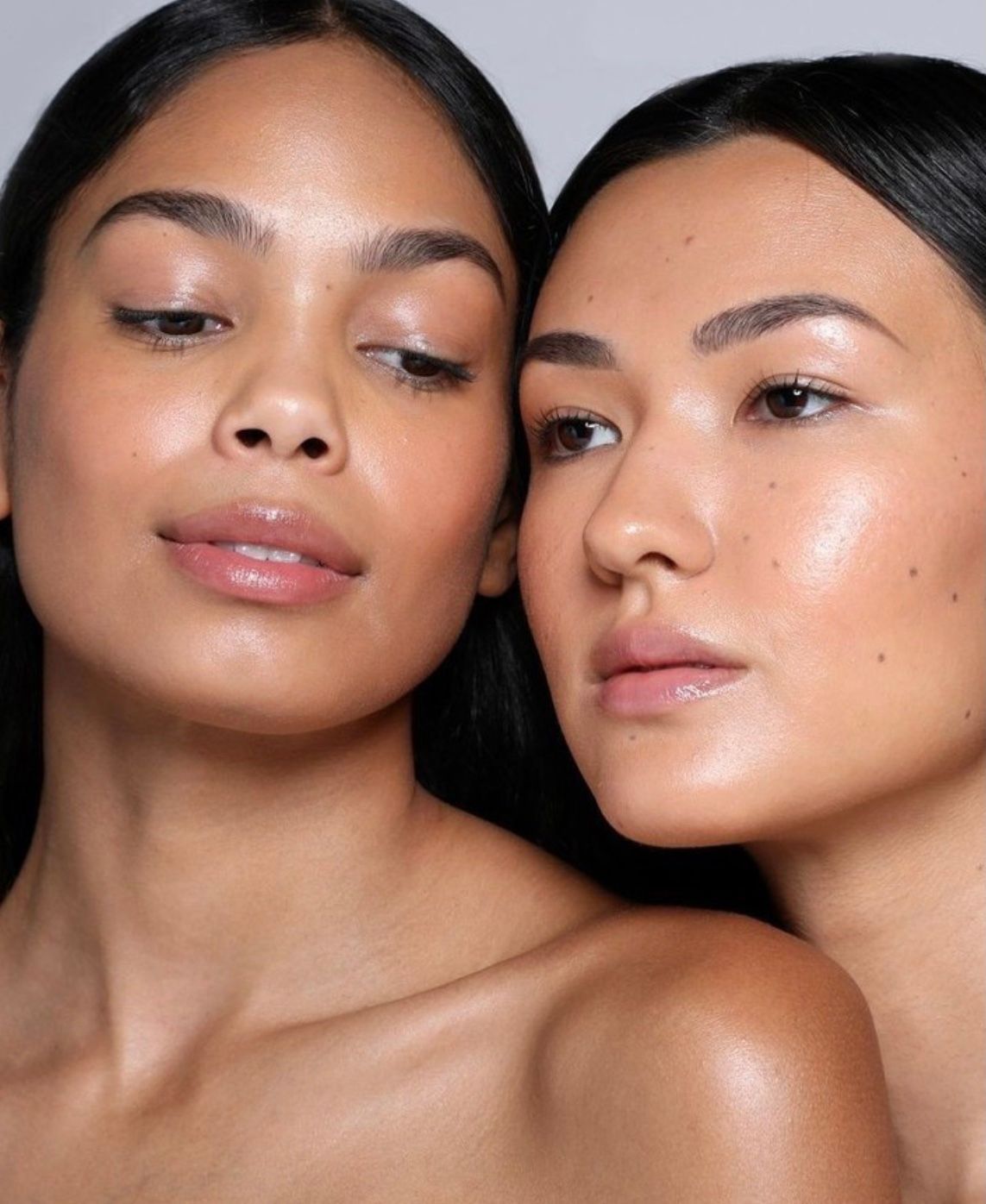 Two women with smooth skin and natural makeup pose closely against a black background.