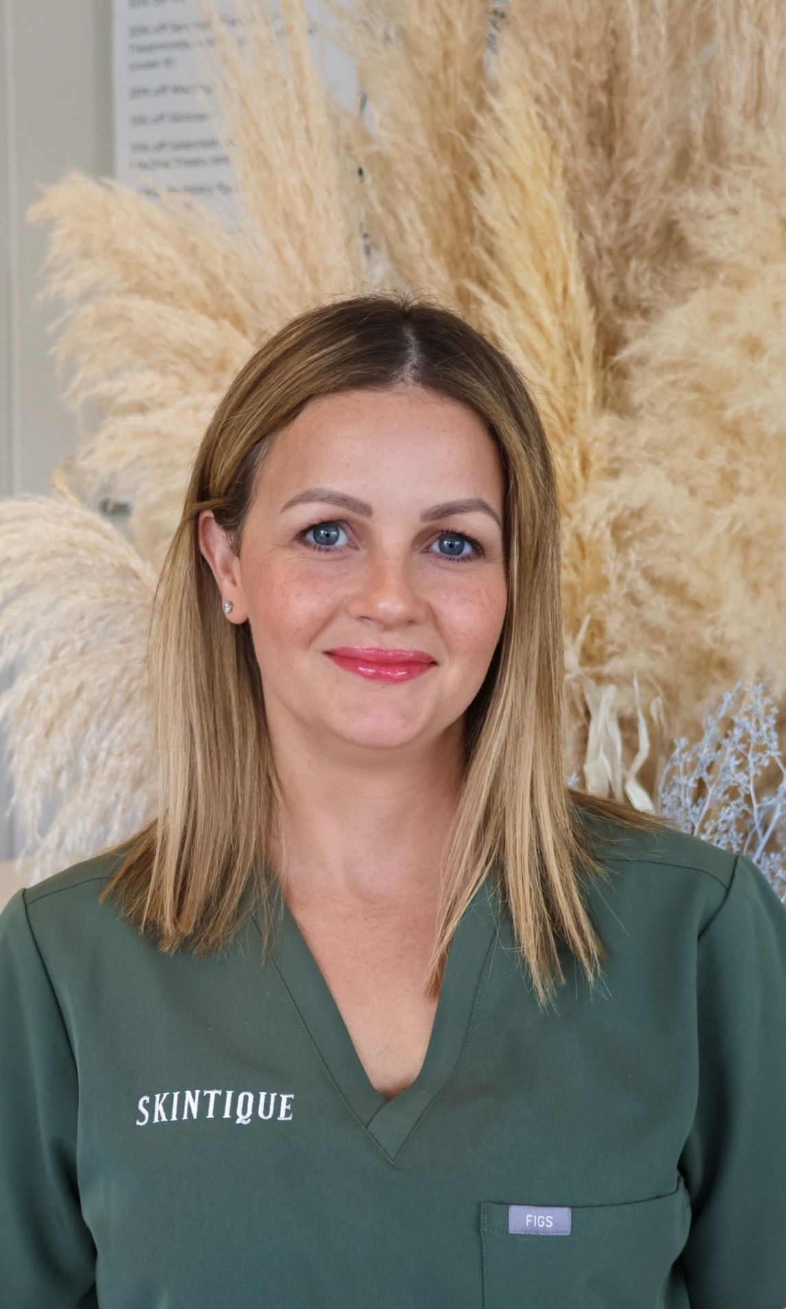 Woman in green top standing before tall beige pampas grass, smiling at the camera.