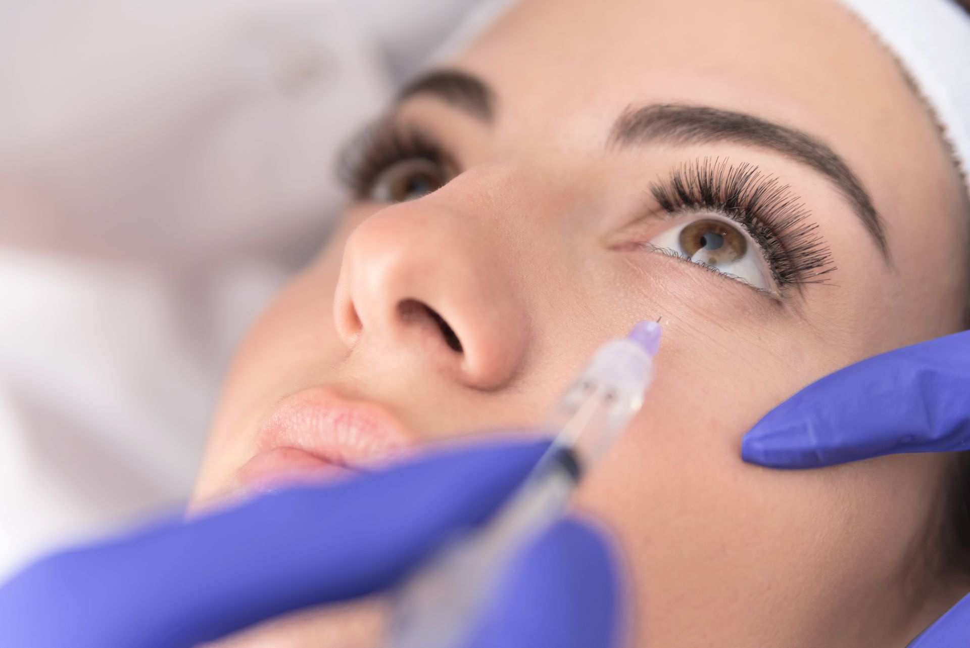 Close-up of a cosmetic injection near a woman’s eye, with gloved hands in a clinical setting