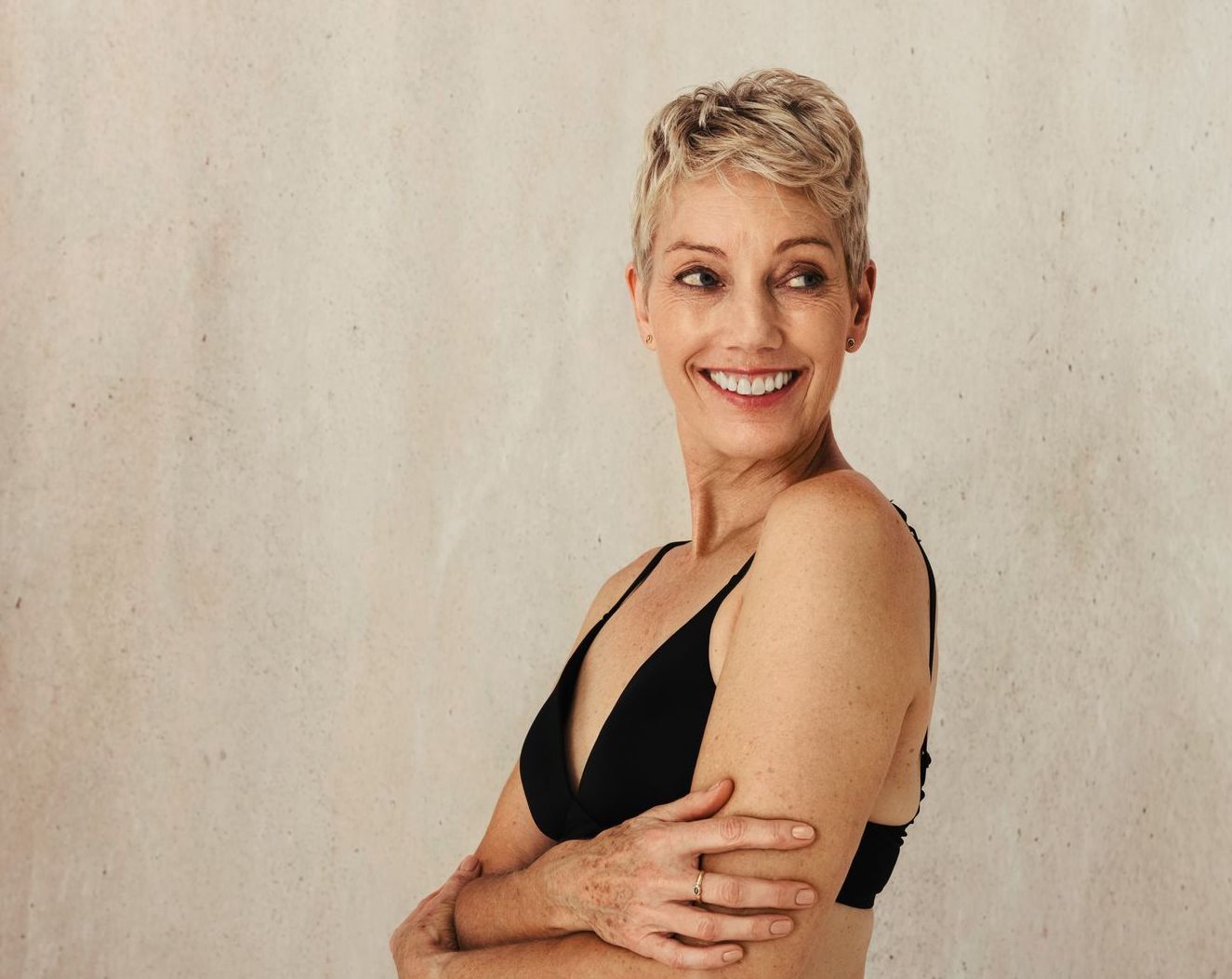 Smiling woman in a black top with crossed arms, standing against a light wall.