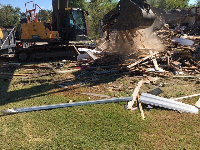 Excavator demolishing a structure, kicking up dust and debris on a grassy area.