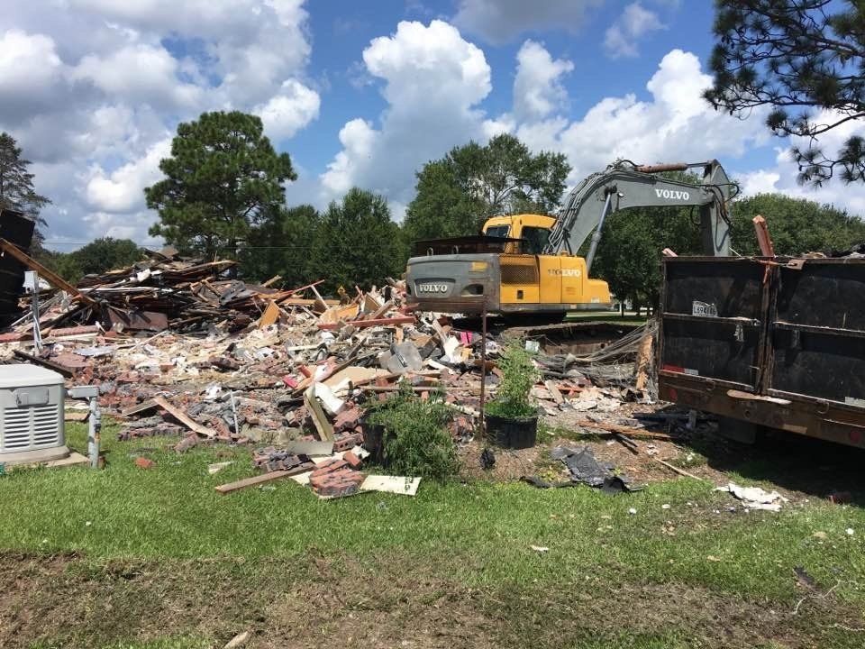 Demolished building with yellow excavator and debris in a yard, under a cloudy blue sky.