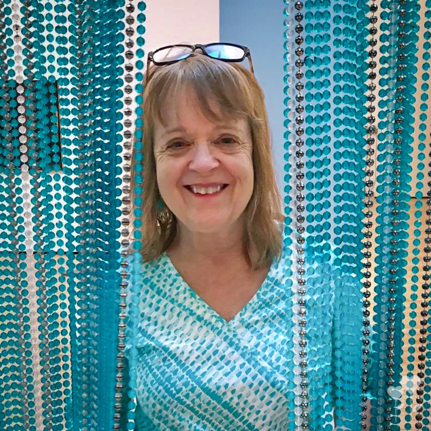 a woman wearing glasses stands in front of a wall of blue beads