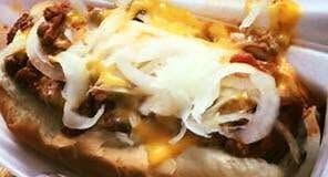 Close-up of a hot dog topped with melted cheese, onions, and chili in a paper tray