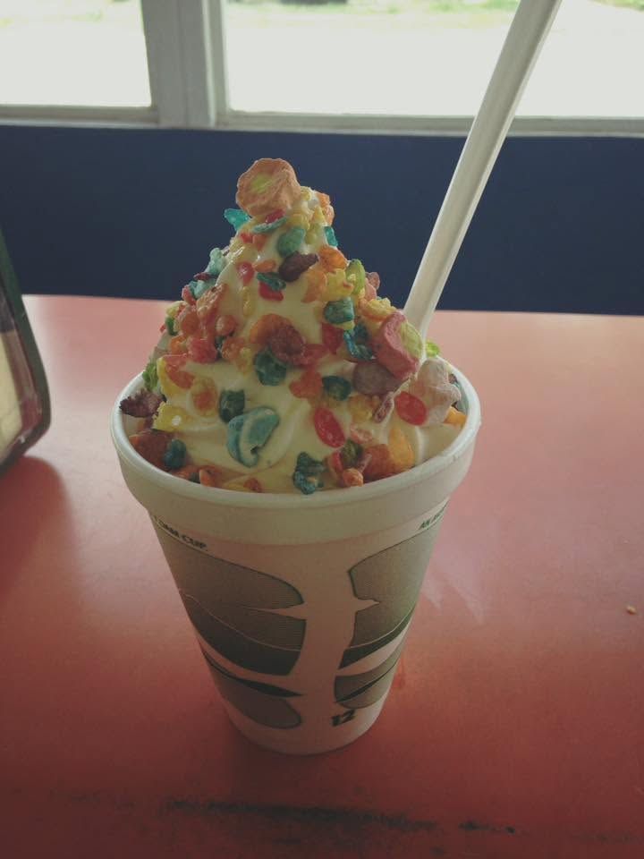 Colorful sprinkle-topped soft serve in a paper cup with a spoon on a table