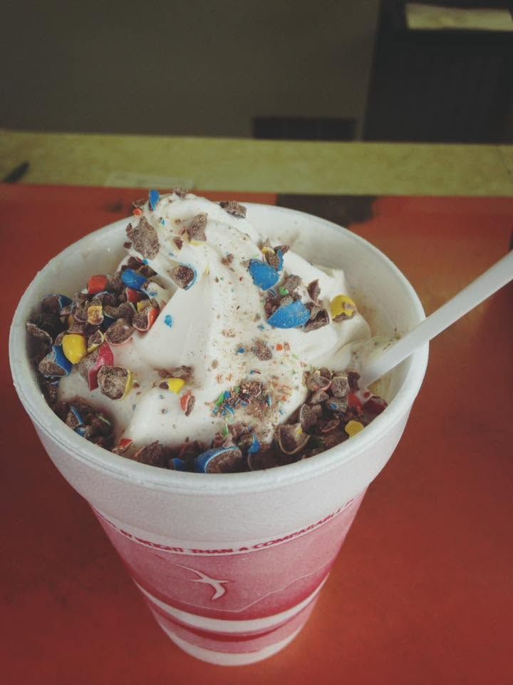 Cup of soft-serve ice cream topped with colorful sprinkles and sauce, with a spoon in a pink cup