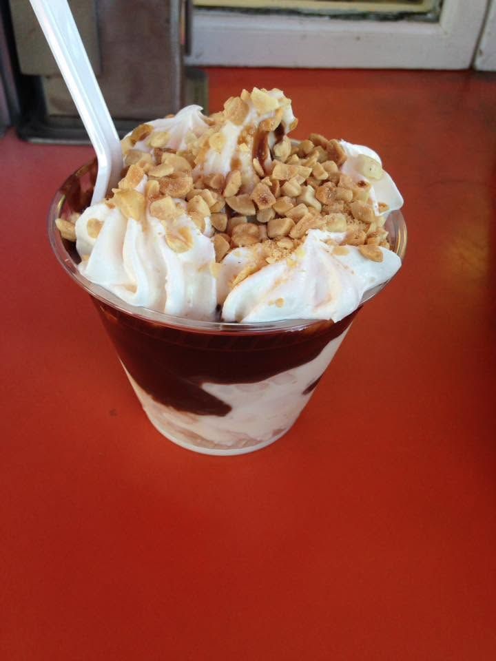 Ice cream sundae with whipped cream, caramel drizzle, and chopped nuts in a clear cup on a red table.