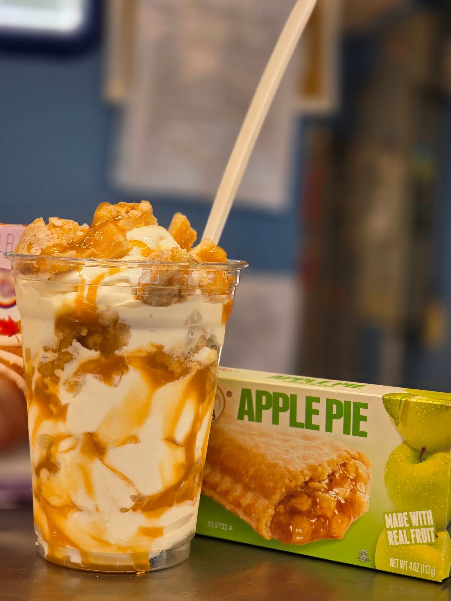 Apple pie sundae in a clear cup with caramel drizzle and a spoon, beside an Apple Pie dessert box.