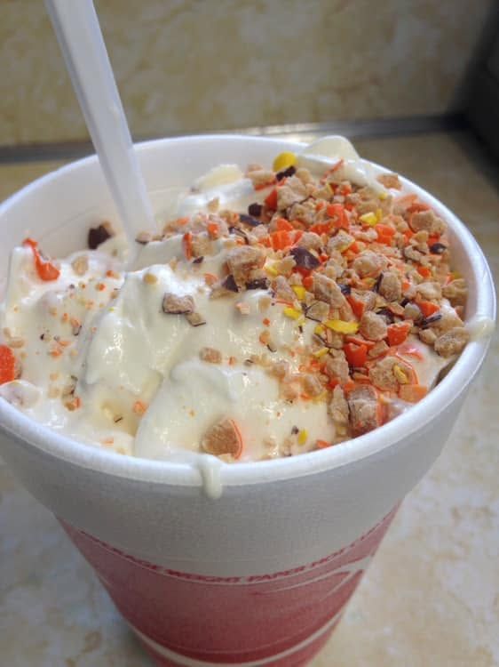 A cup of ice cream topped with crushed nuts and candy bits, with a spoon inside.