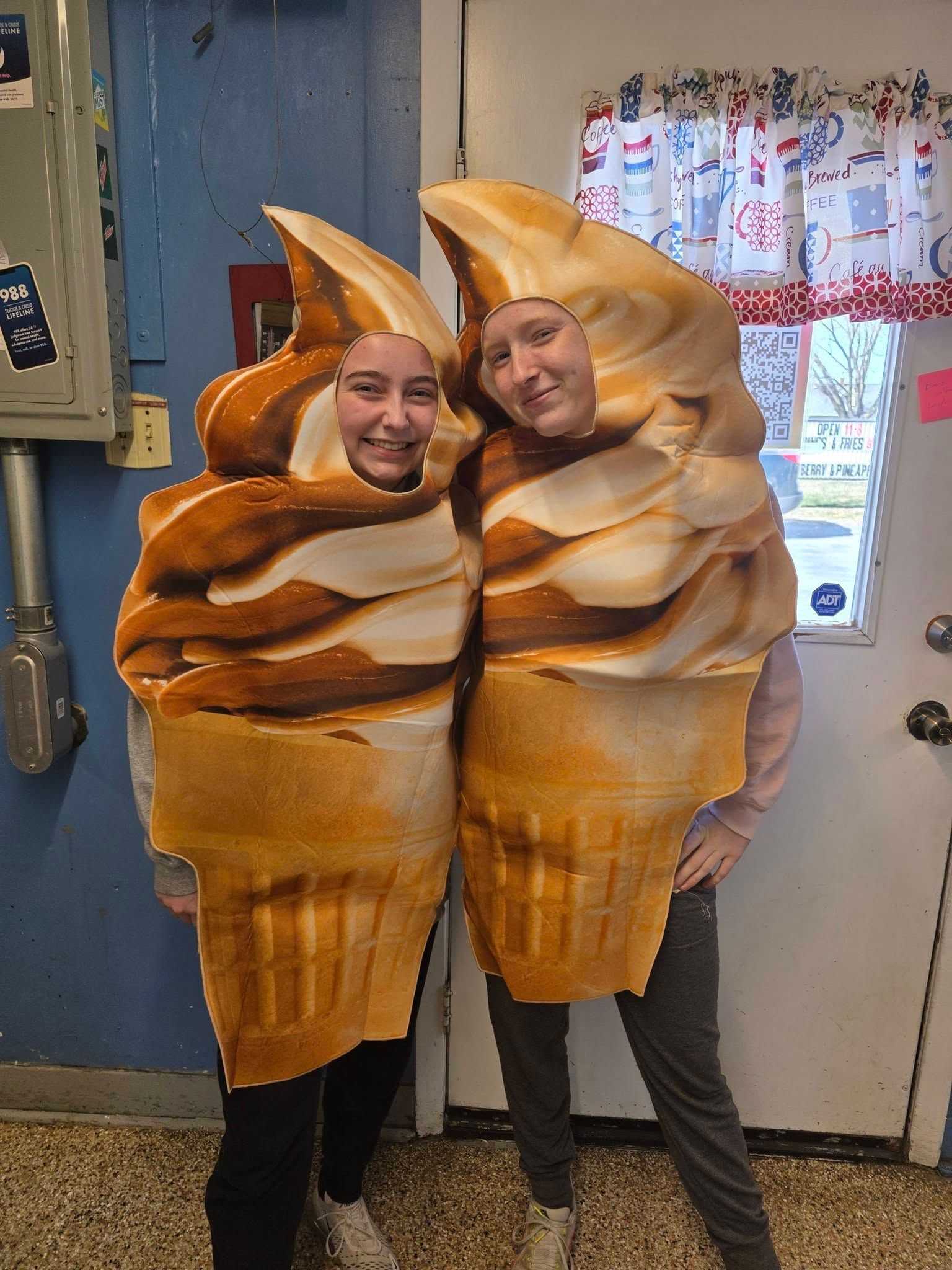 Two people in pastry costumes smiling and standing together indoors by a door and window.