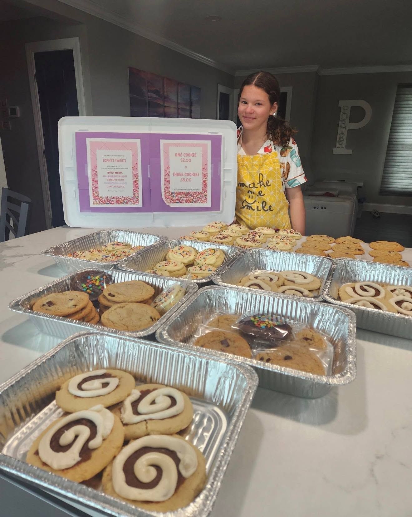 Woman stands behind a table of assorted baked goods with a framed sign in a home kitchen.