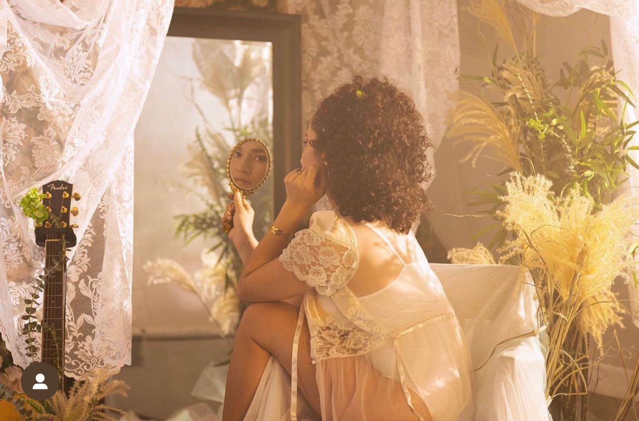 Woman siting in sunlit room wearing white dress staring into hand mirror