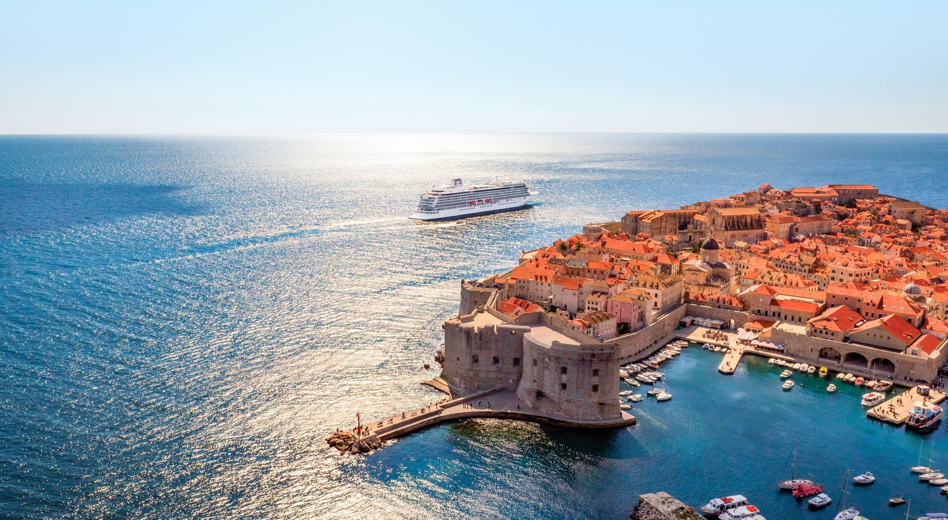 Coastal city with orange rooftops, stone walls, and boats, a large cruise ship in the distance under a bright blue sky.
