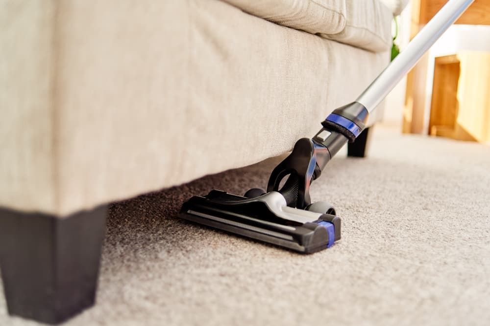 Vacuuming carpet under a beige sofa with a handheld vacuum tool