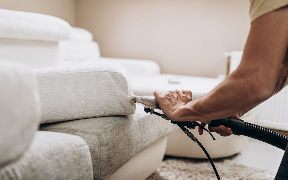 Person vacuuming a white sofa with a handheld upholstery cleaner in a living room