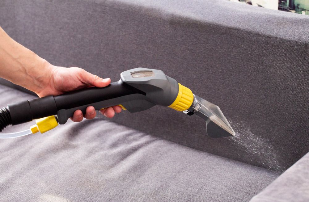 Handheld upholstery cleaner spraying water on a gray sofa cushion