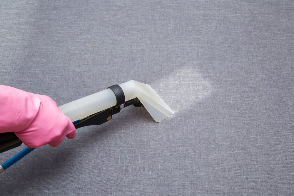 A gloved hand using a vacuum cleaner nozzle on a gray upholstered surface