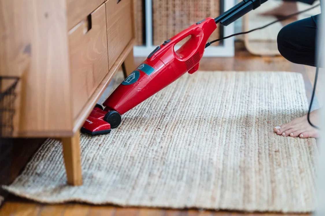 Red vacuum cleaner on a beige rug beside wooden furniture, with a person’s bare feet nearby.