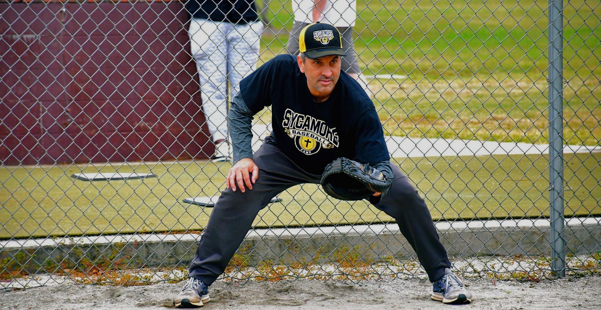 Man in baseball gear fielding on a field, wearing a cap, dark shirt, and gray pants.