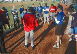 Group of people in athletic gear, standing in a circle on a dirt field, having a prayer.