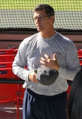 Man in grey long-sleeve shirt and glasses holds a hat, standing near a baseball dugout.