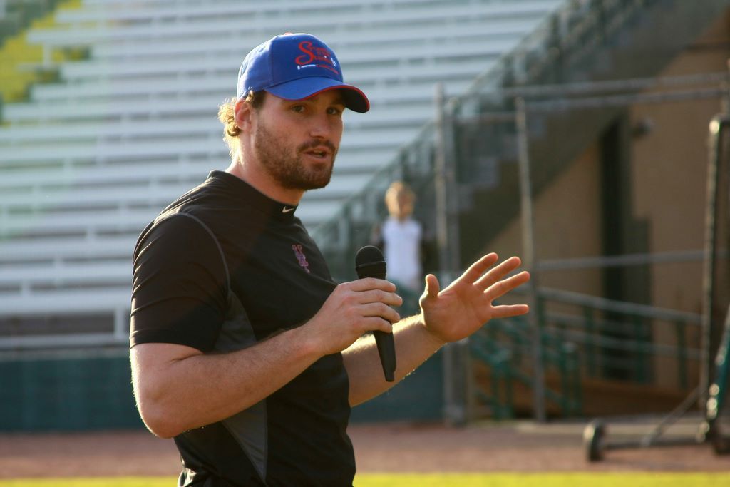 Man in baseball cap holding microphone, gesturing on field.