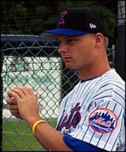 Baseball player in a Mets uniform holding a ball, wearing a cap, standing near a fence.
