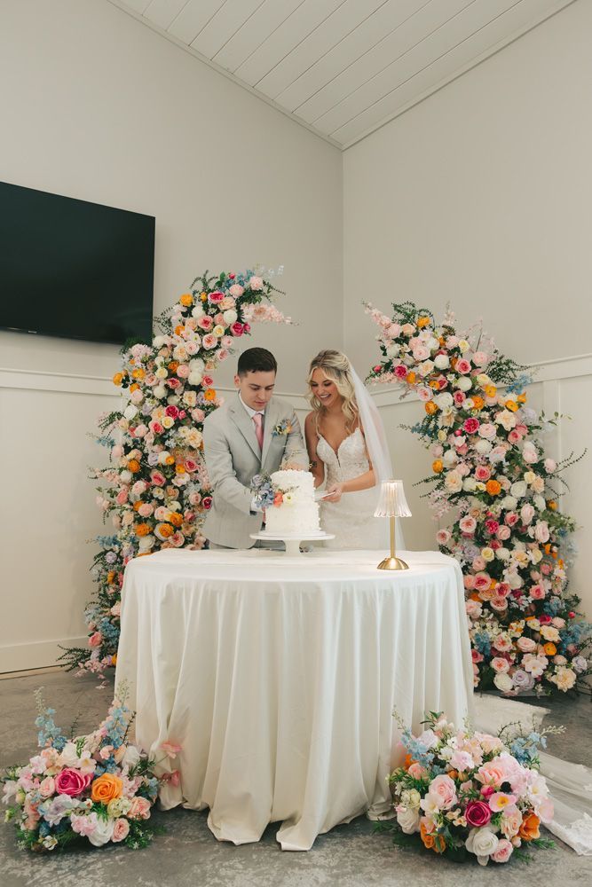 Couple cutting a wedding cake beneath a floral arch in a bright indoor ceremony setting