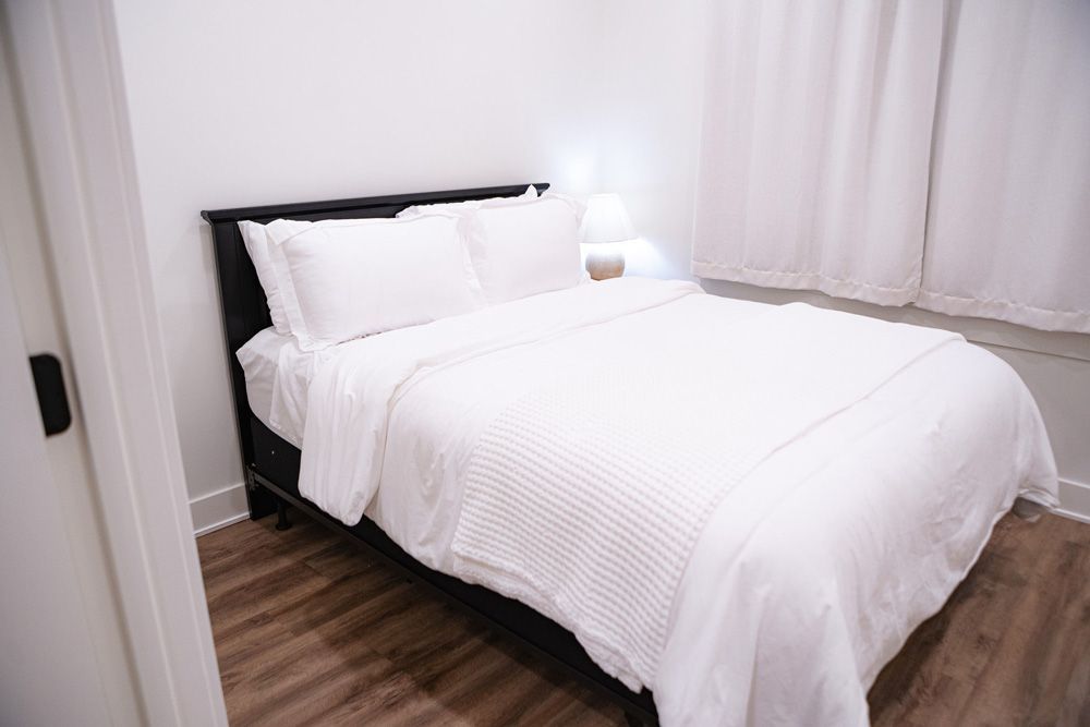 Small bedroom with a neatly made white bed, bedside lamp, and wood floor.