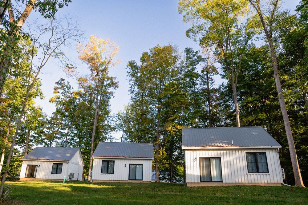 Three small white cabins with dark roofs in a grassy clearing, backed by tall autumn trees under a blue sky