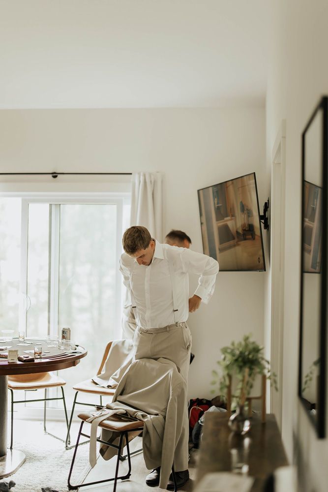 Man in white shirt adjusting pants in a bright dining room with chairs and a table by a large window