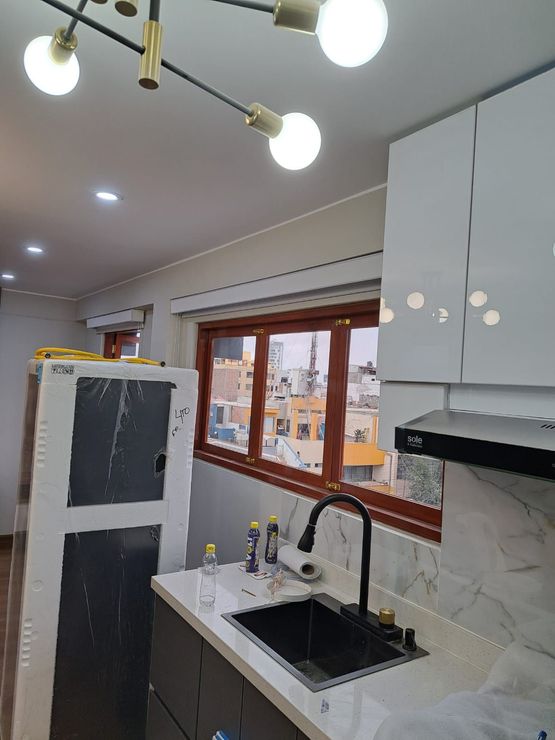 Tres pintores vestidos de azul y naranja trabajan en la pared de una habitación luminosa con rodillos de pintura y lonas protectoras.
